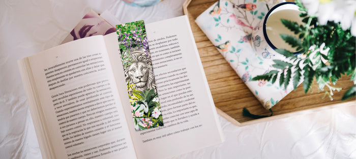 Marque-page printanier représentant un lion entouré de fleurs et de végétation, posé dans un livre ouvert avec une tasse de thé et des plantes sur une table